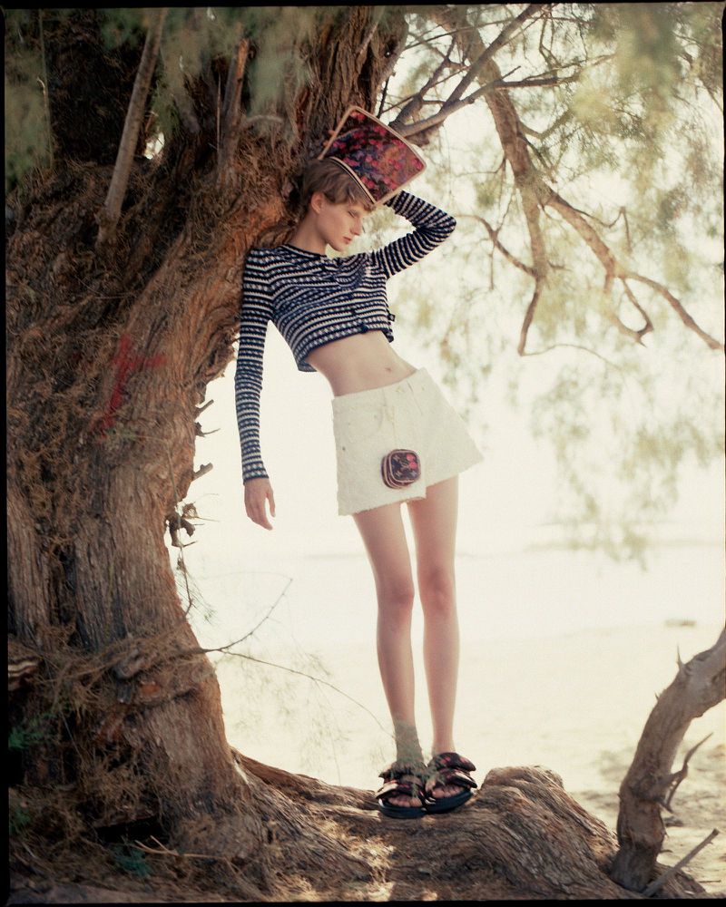 A slender fashion model poses near a tree on a beach wearing a striped top, white skirt, socks, sandals, and holding a magazine.