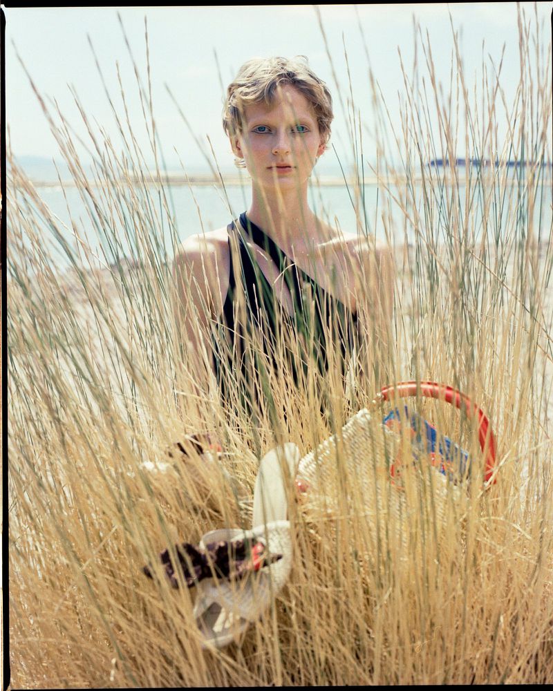 A woman in a black top stands amidst tall, tan beach grass near a lake, holding a handbag and shoe in a sunlit outdoor setting.
