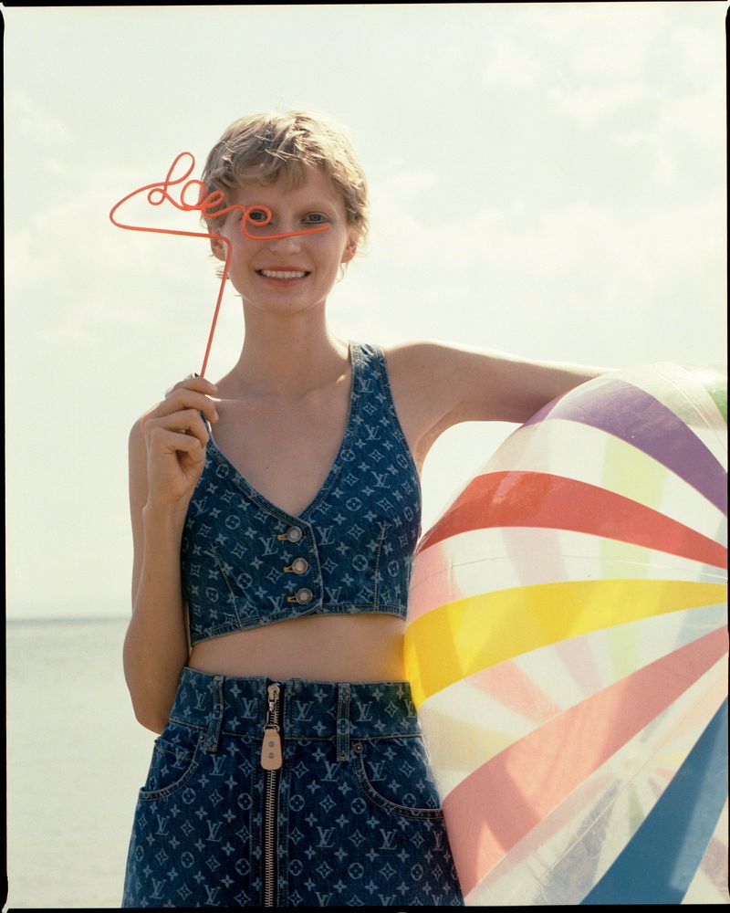 Blonde woman on beach smiles holding 'love' sign, wearing designer denim outfit and next to colorful beach ball. Summery, bright setting.