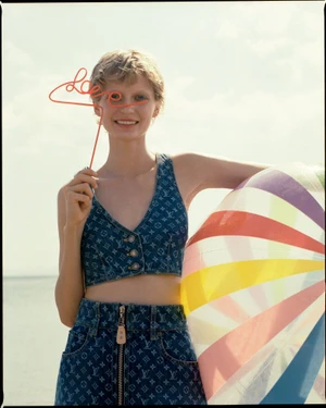 Model in monogram denim crop top holding a red wire prop and striped beach ball against an ocean backdrop.