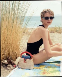 Model in black swimsuit and sunglasses seated on a beach with a woven handbag and sandals on a map