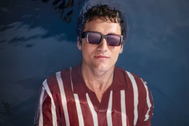 Man wearing dark sunglasses and a red and white striped polo shirt submerged in water up to his chest, captured by Seiya Taguchi for Specsavers.