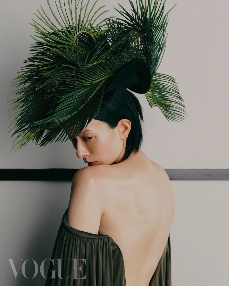 An adult female with short black hair looks to the side, wearing an open-back draped dress and elaborate leafy headdress in a neutral studio.