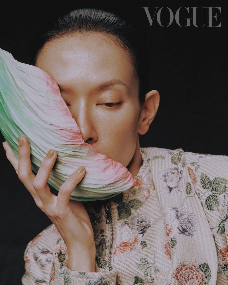 Close-up fashion portrait of an adult woman with short hair in a floral jacket holding a folded, fan-like prop against a dark background.