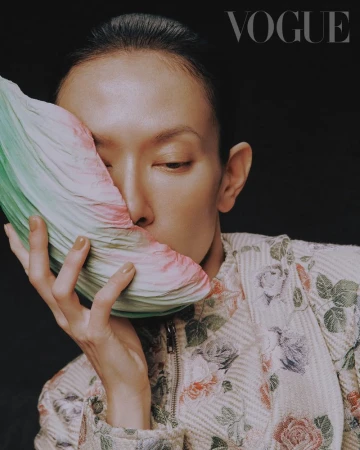Close-up fashion portrait of an adult woman with short hair in a floral jacket holding a folded, fan-like prop against a dark background.
