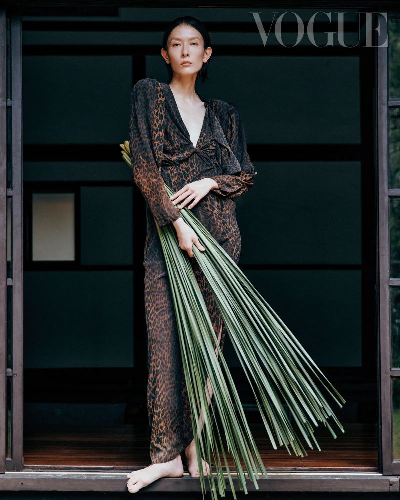 A woman in an animal-print dress stands in a doorway holding foliage, featured in Vogue, showcasing fashion and photographic artistry.