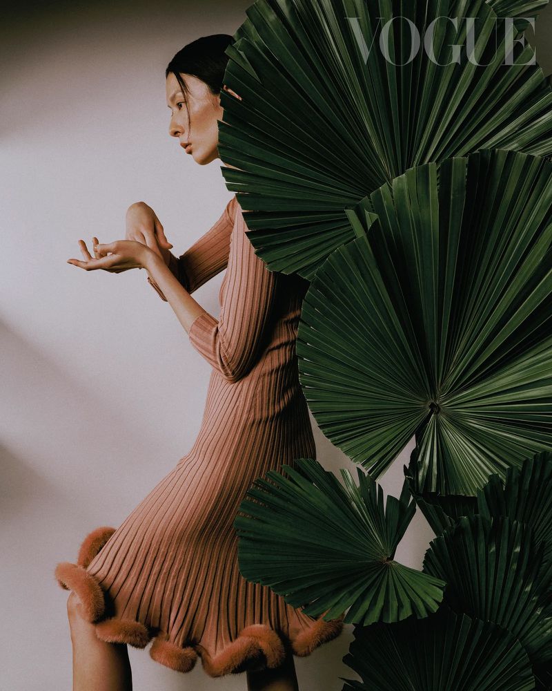 Fashion editorial shot of a woman in a pink ribbed dress posing near large green palm leaves against a pale backdrop. Vogue magazine logo visible.