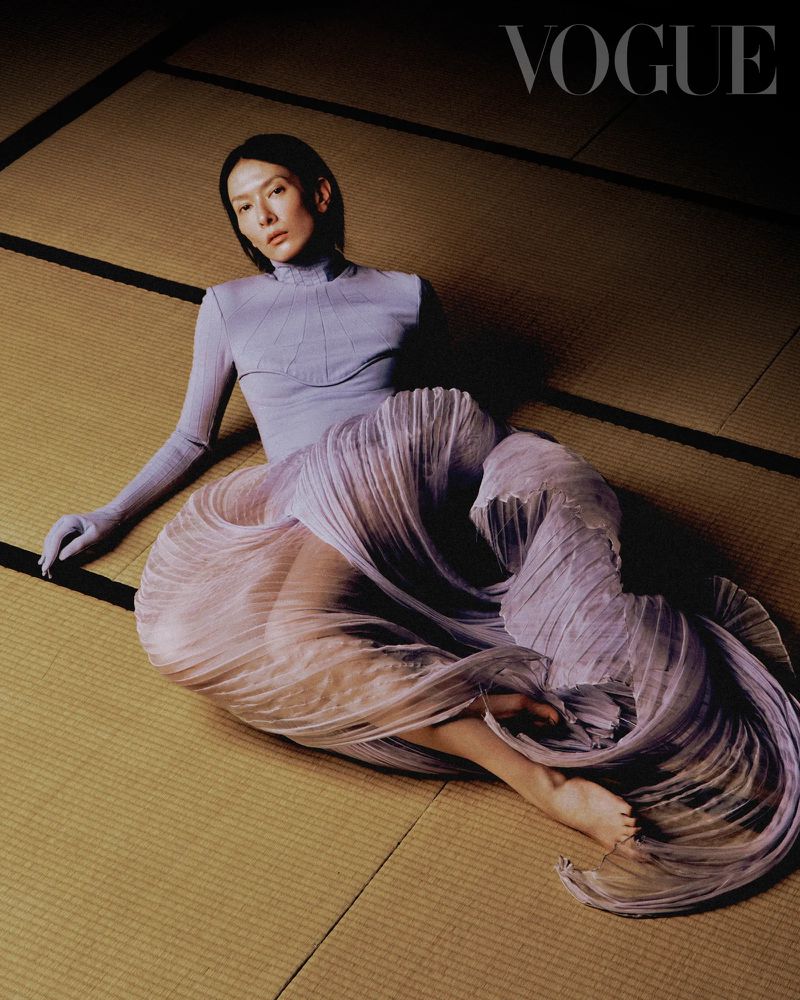 Elegant woman in a light purple outfit poses on a tatami mat for a fashion editorial, highlighted by soft lighting and a Vogue logo.