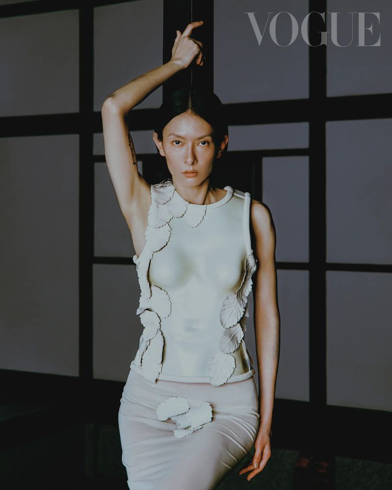 Fashion editorial shot of a slender model in a light-colored top and skirt, posing against a gray and black wall with the VOGUE logo.