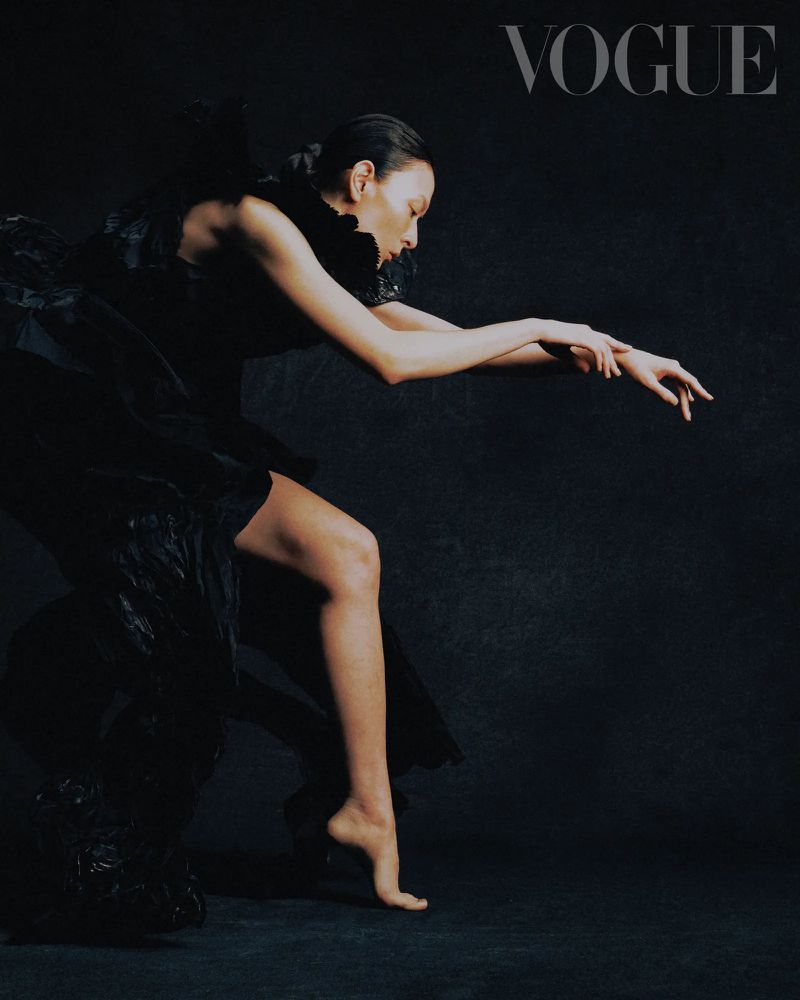 A Vogue cover image features a slender woman in a black ruffled dress, gracefully posing in profile against a dark, monochromatic background.