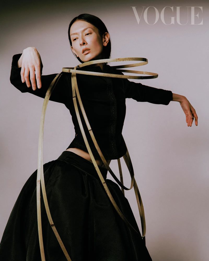 Fashion editorial shot: A slender woman with short dark hair poses in a black dress with decorative beige hoops in a photo studio.