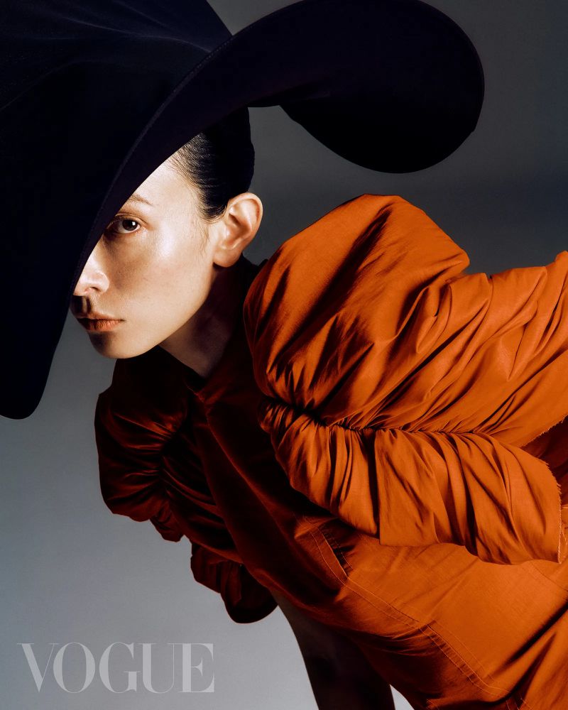 Fashion editorial portrait of a woman wearing an orange dress and a large black hat in front of a grey background with the Vogue logo.