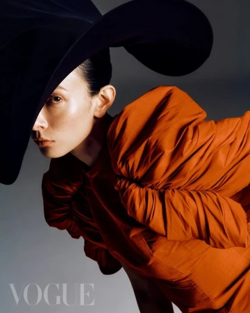 Fashion editorial portrait of a woman wearing an orange dress and a large black hat in front of a grey background with the Vogue logo.