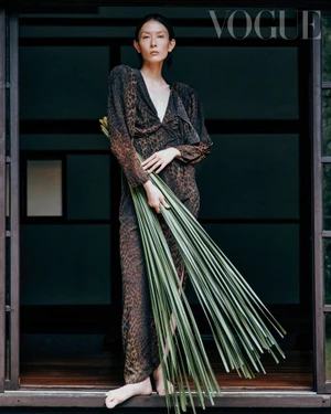 Model Hsin-Yu Liu poses barefoot in a leopard-print dress holding a bundle of green palm fronds in a dark timber doorway.