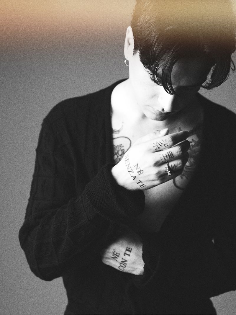Adult male with tattoos poses indoors, wearing a cardigan. Black and white studio-style portrait with a shallow depth of field.