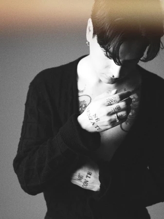 Adult male with tattoos poses indoors, wearing a cardigan. Black and white studio-style portrait with a shallow depth of field.