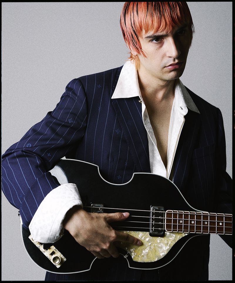 A man with pink hair wearing a pinstripe suit poses indoors with a black hollow body guitar against a neutral grey backdrop.