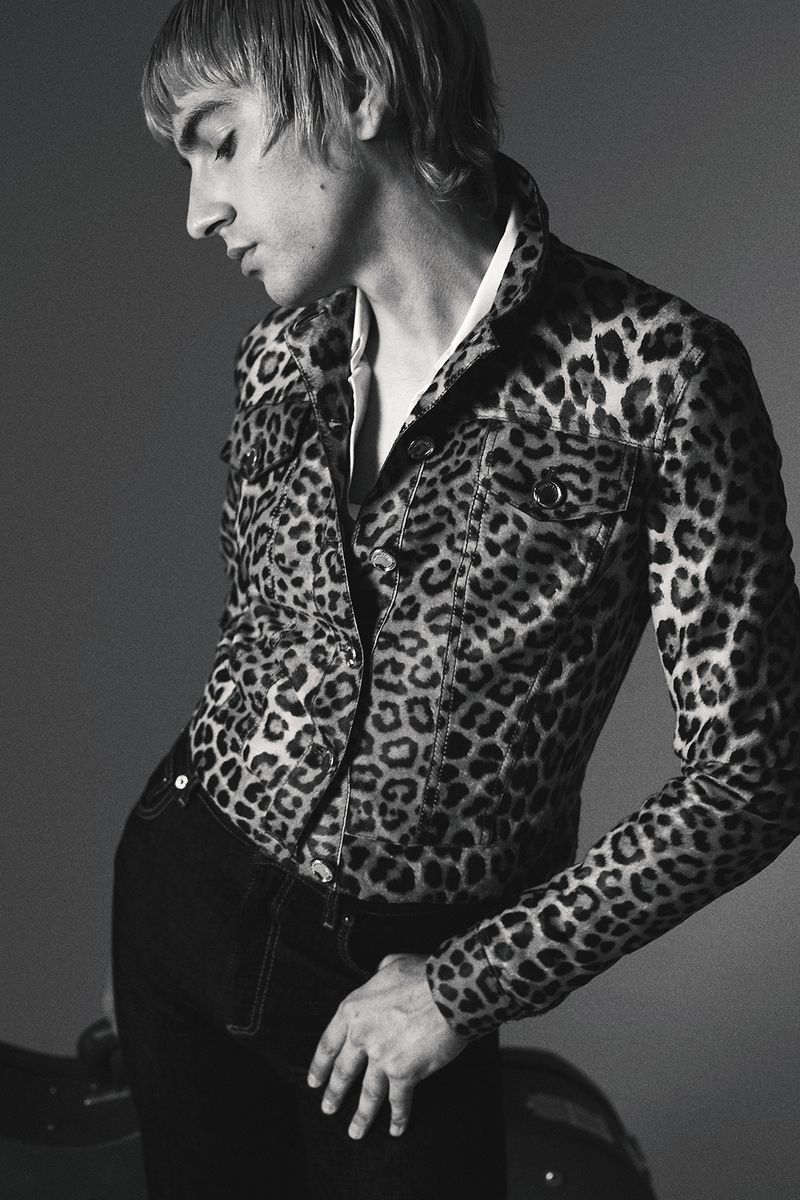 Black and white portrait of a slender male in an open leopard print jacket, wearing dark jeans, in a studio setting, with a reflective prop.