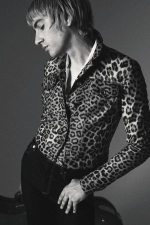 Black and white portrait of a slender male in an open leopard print jacket, wearing dark jeans, in a studio setting, with a reflective prop.