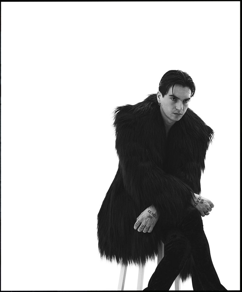 Portrait of a man in a dark fur coat sitting on a stool against a white background in a studio, black and white fashion shot.