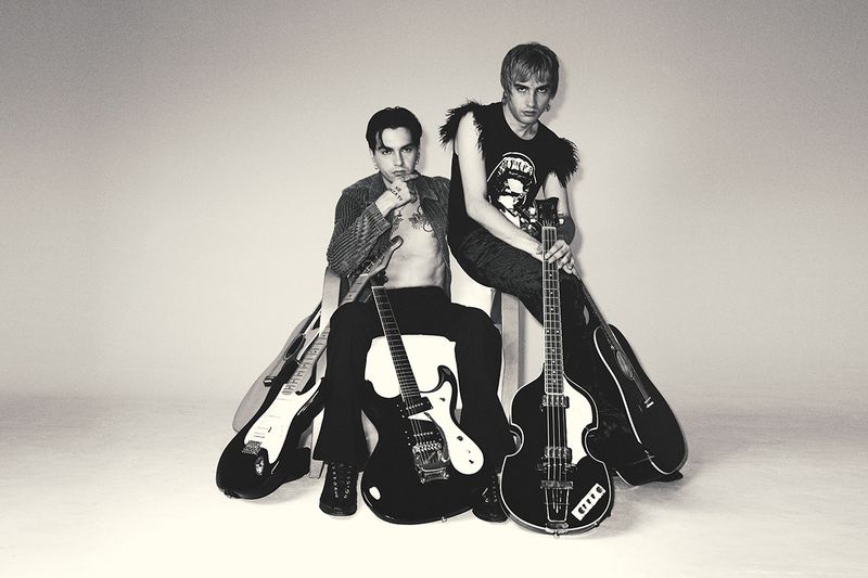 Two musicians, one seated and bare-chested wearing black pants and a striped jacket, and another seated with a graphic tee and feather boa, each holding guitars.