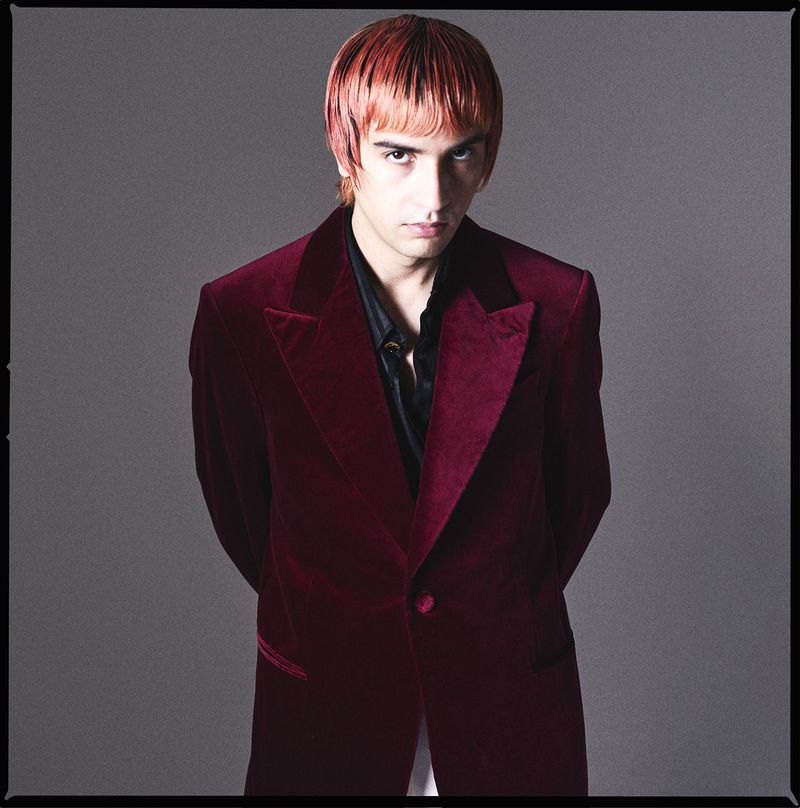 Portrait of a young adult male with dyed orange hair, wearing a deep red velvet suit jacket and a dark collared shirt against a gray background.