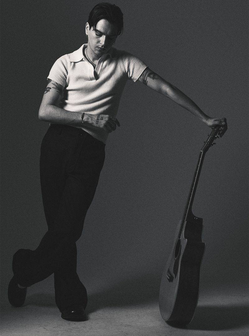Monochrome portrait of a man with tattoos leaning on a guitar, wearing a collared shirt and dark pants, standing indoors against a grey backdrop.