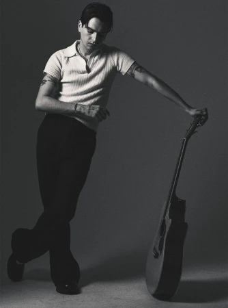Monochrome portrait of a man with tattoos leaning on a guitar, wearing a collared shirt and dark pants, standing indoors against a grey backdrop.
