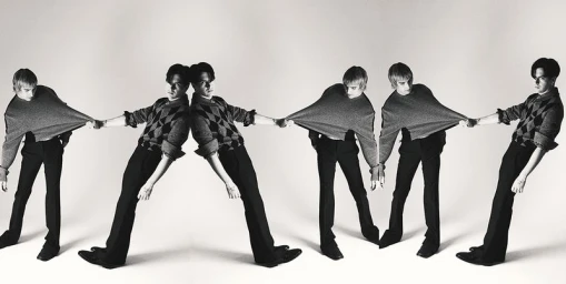 Two male models posing in dynamic pulling gestures wearing knit sweaters and black trousers against a white studio background.