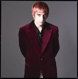 Man wearing a burgundy velvet blazer over a dark shirt against a neutral grey studio background.