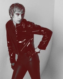Man wearing a dark glossy jacket and tight trousers posing against a neutral textured studio background.