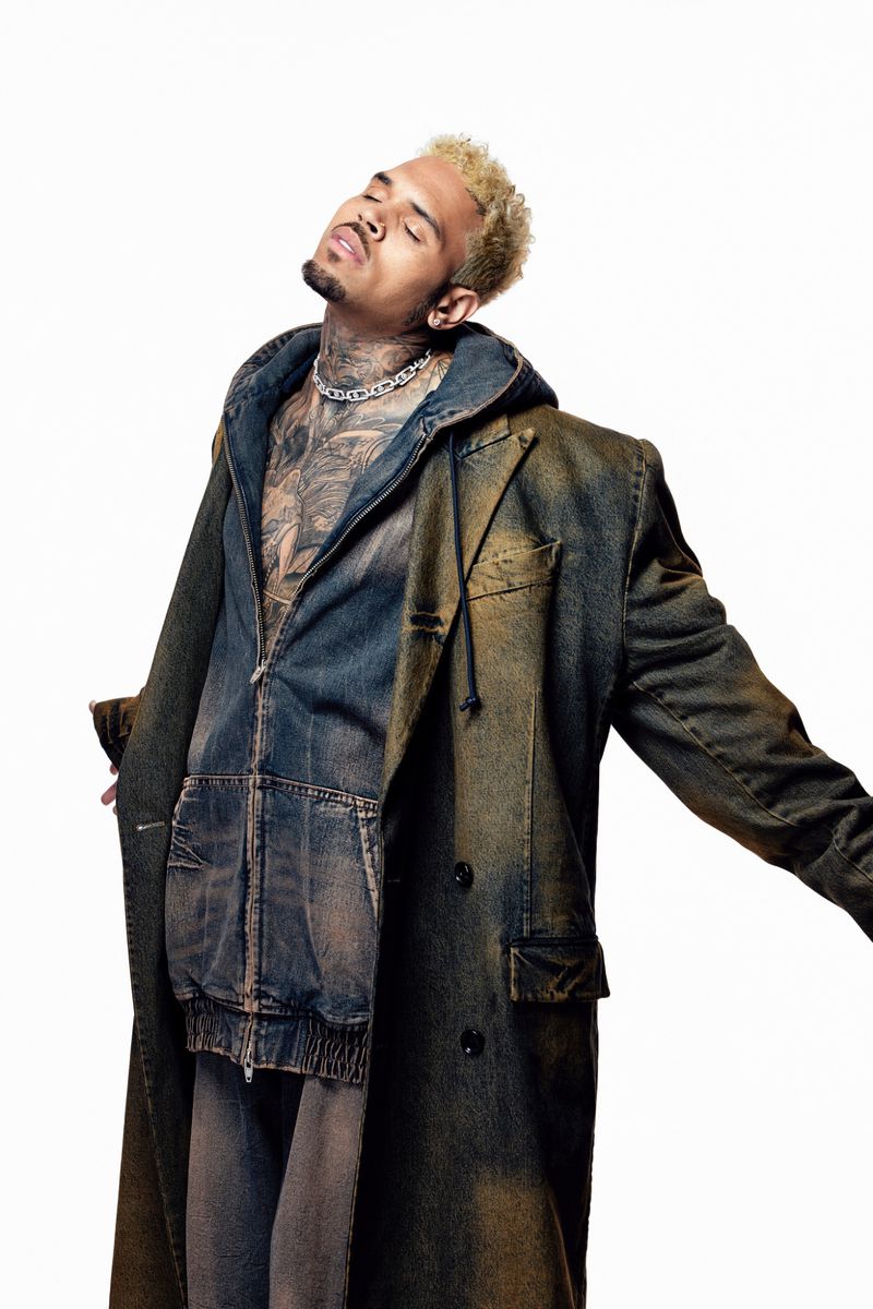 Chris Brown poses in a denim ensemble, featuring a long coat and hooded jacket, against a clean white backdrop in a studio setting.