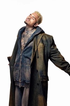 Chris Brown poses in a denim ensemble, featuring a long coat and hooded jacket, against a clean white backdrop in a studio setting.