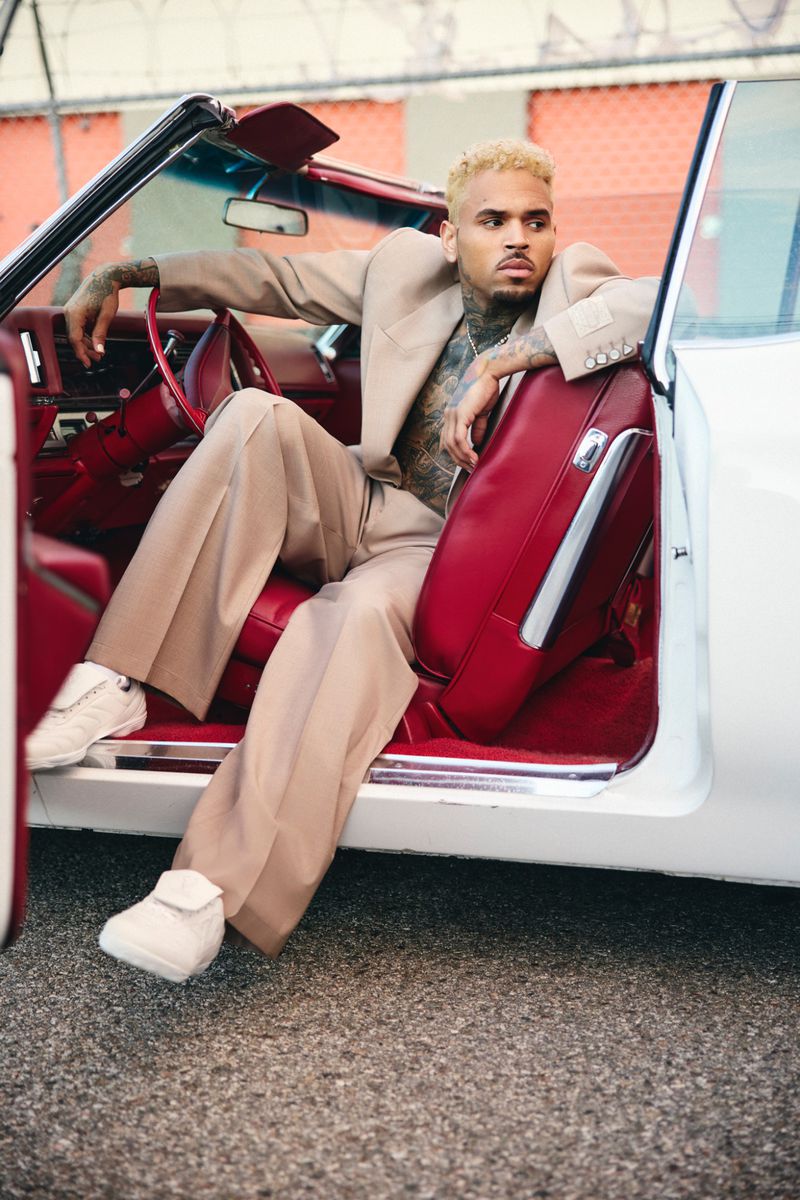 Adult male poses in white convertible car wearing tan suit and white sneakers, looking off camera with building in the background.