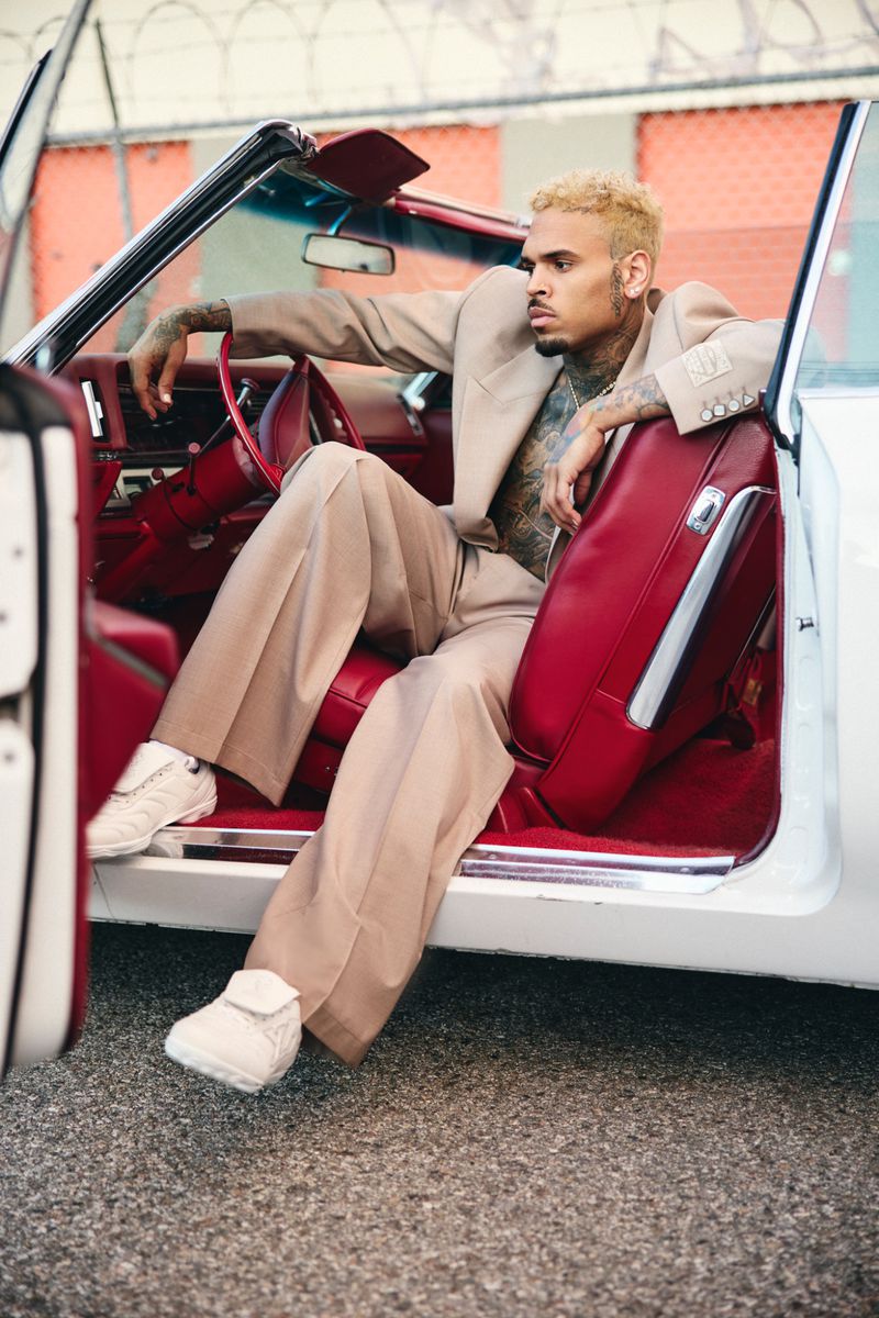 Chris Brown poses inside a vintage car wearing a suit with open door. The setting is an urban street with natural light.