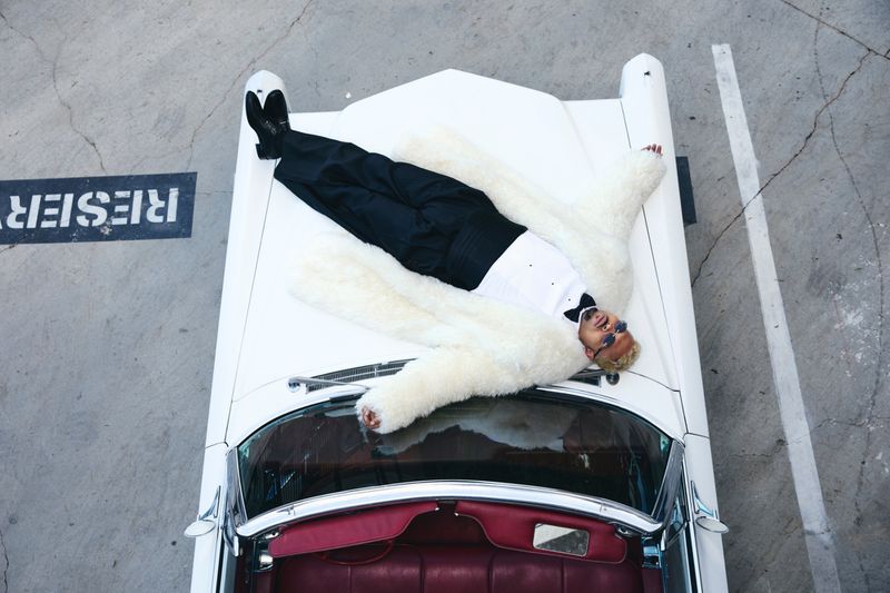 Overhead shot of a blond adult male in a white tuxedo shirt and fur coat reclining on the hood of a white classic car in parking lot.