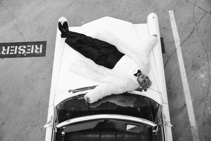 Black and white shot of a male lying on the hood of a white classic car wearing a tuxedo and a fur coat in an indoor environment.