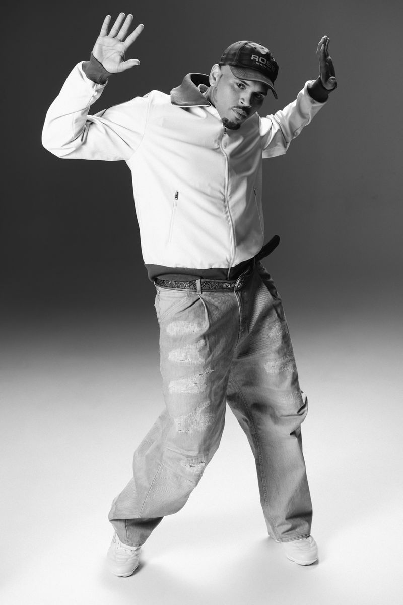 Black and white studio shot of an adult male in a light jacket, baggy jeans, and a baseball cap with his hands raised up.
