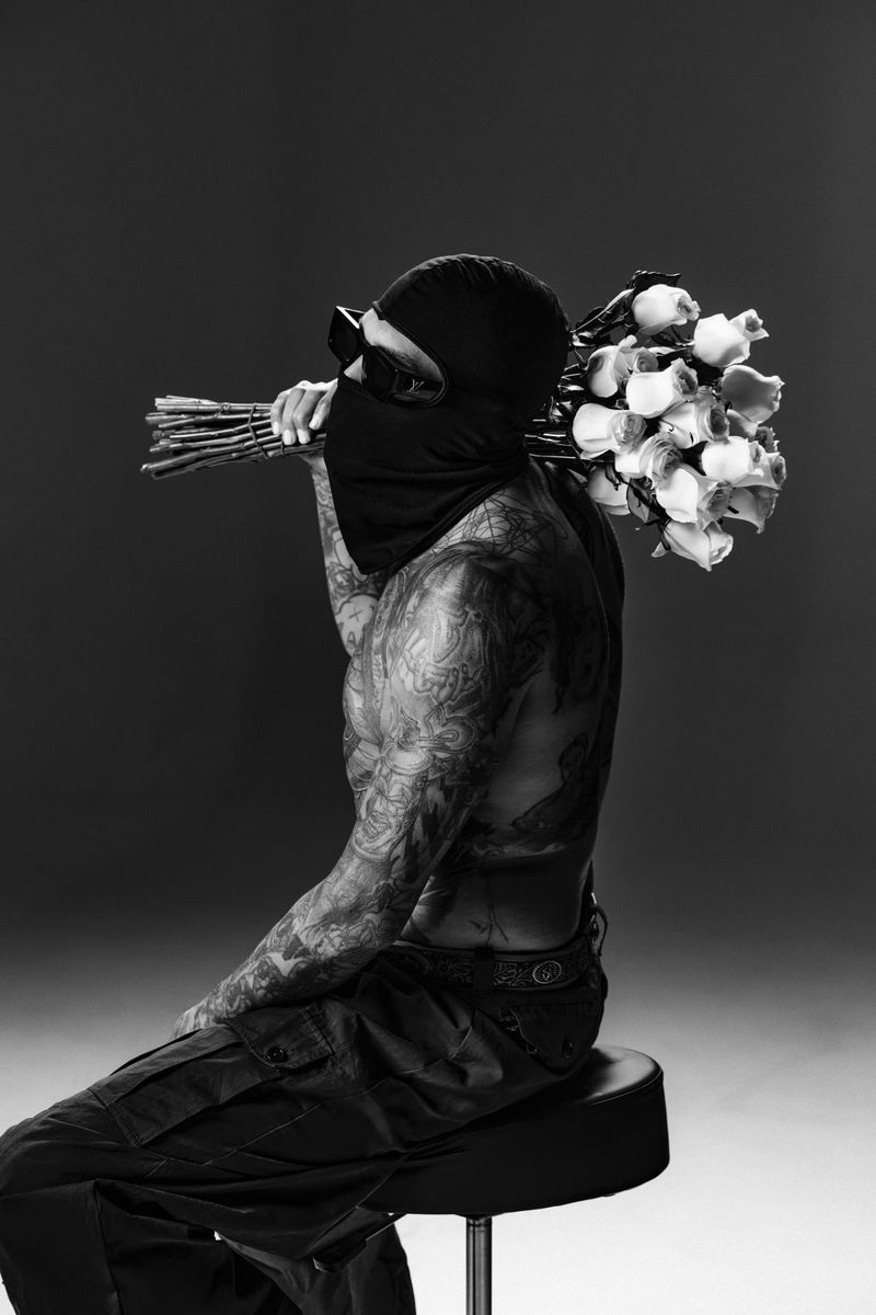 Monochrome portrait of tattooed man in ski mask sitting on stool and holding bouquet of roses over shoulder in studio setting.