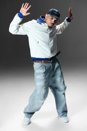 Full-length portrait of a man in a light jacket and baggy jeans striking a hip-hop dance pose against a gradient gray background.