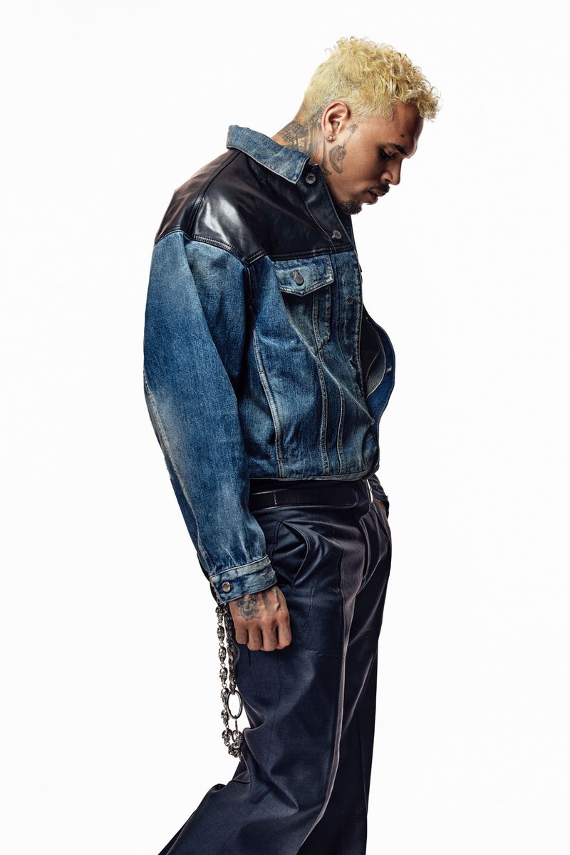 Side profile of a man with bleached blonde hair, wearing a two-tone denim jacket, and black trousers against a bright white backdrop.
