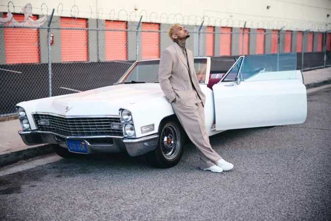 Chris Brown poses against a white Cadillac convertible in an urban setting, wearing a stylish tan suit and white sneakers, looking upwards.
