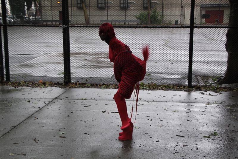 An adult person poses in a vibrant red knitted outfit and chunky heels on a wet, grey outdoor court with a chain-link fence.