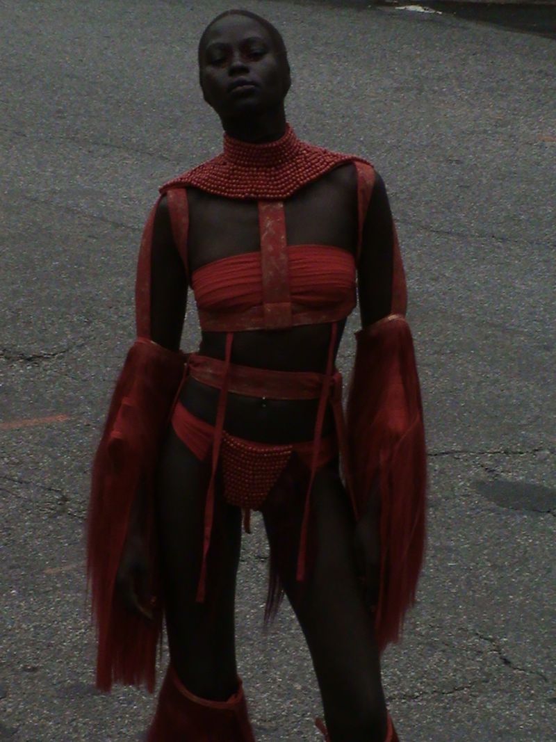 A dark-skinned woman poses in a red, semi-sheer beaded and fur-trimmed outfit against a gray urban background for a fashion shot.