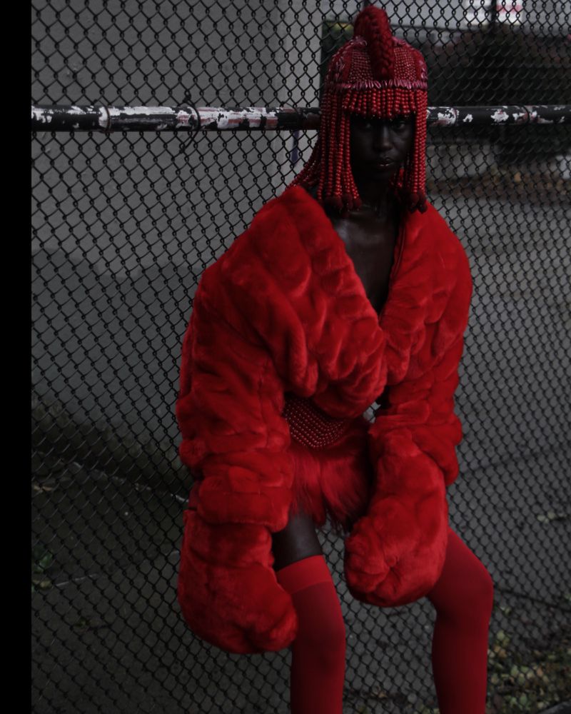 A person in a red beaded helmet and a red faux-fur coat poses in front of a fence, creating a striking and textured fashion image.