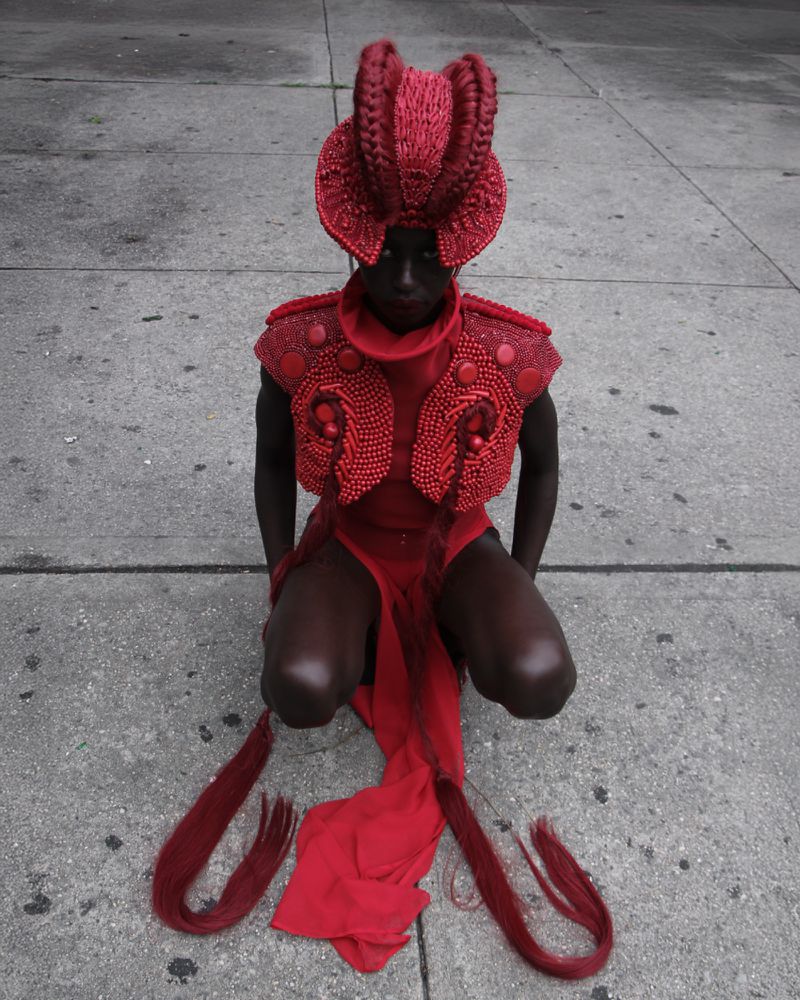 Person in red headpiece and clothing kneels on concrete, facing forward. High-fashion editorial shot. Dark skin, street setting.