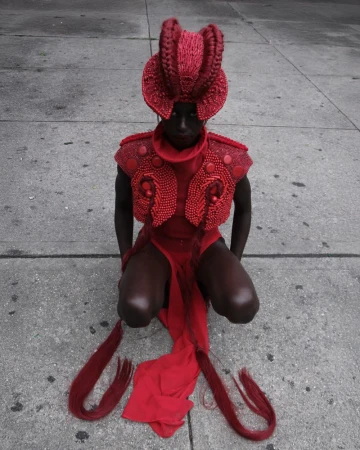 Person in red headpiece and clothing kneels on concrete, facing forward. High-fashion editorial shot. Dark skin, street setting.