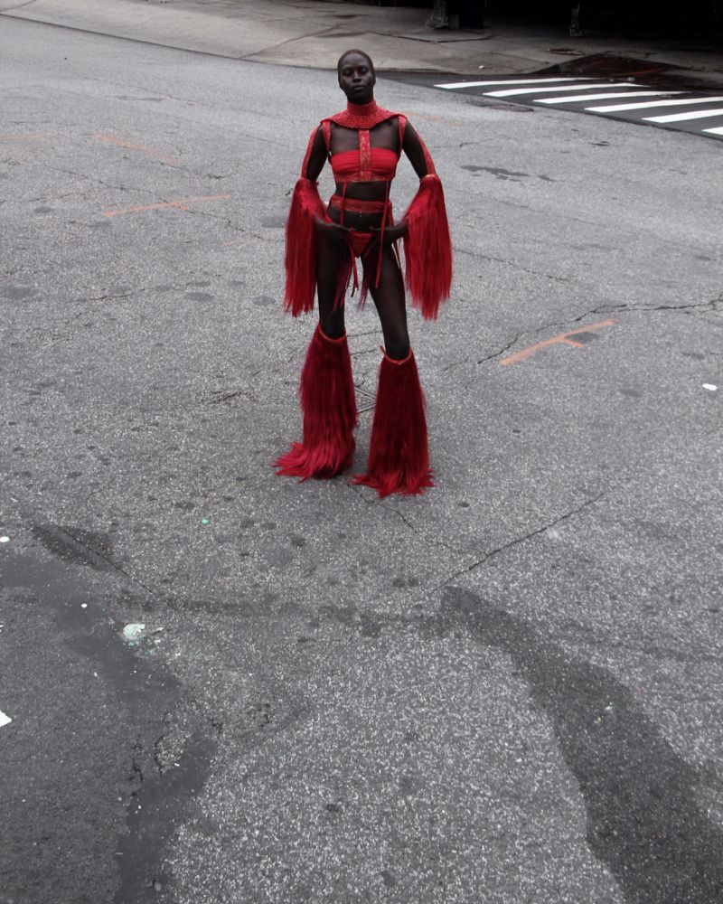 Woman in red costume with fur accents poses on an asphalt road with pedestrian crossing in the background. Full body shot, urban scene.
