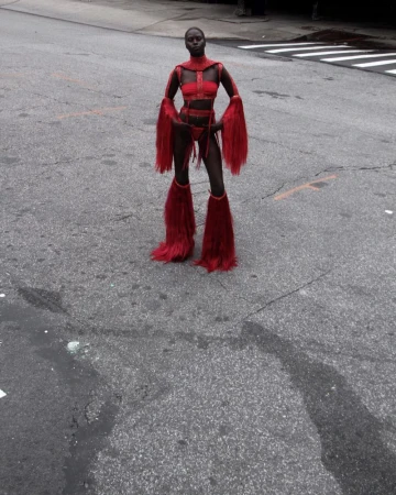 Woman in red costume with fur accents poses on an asphalt road with pedestrian crossing in the background. Full body shot, urban scene.