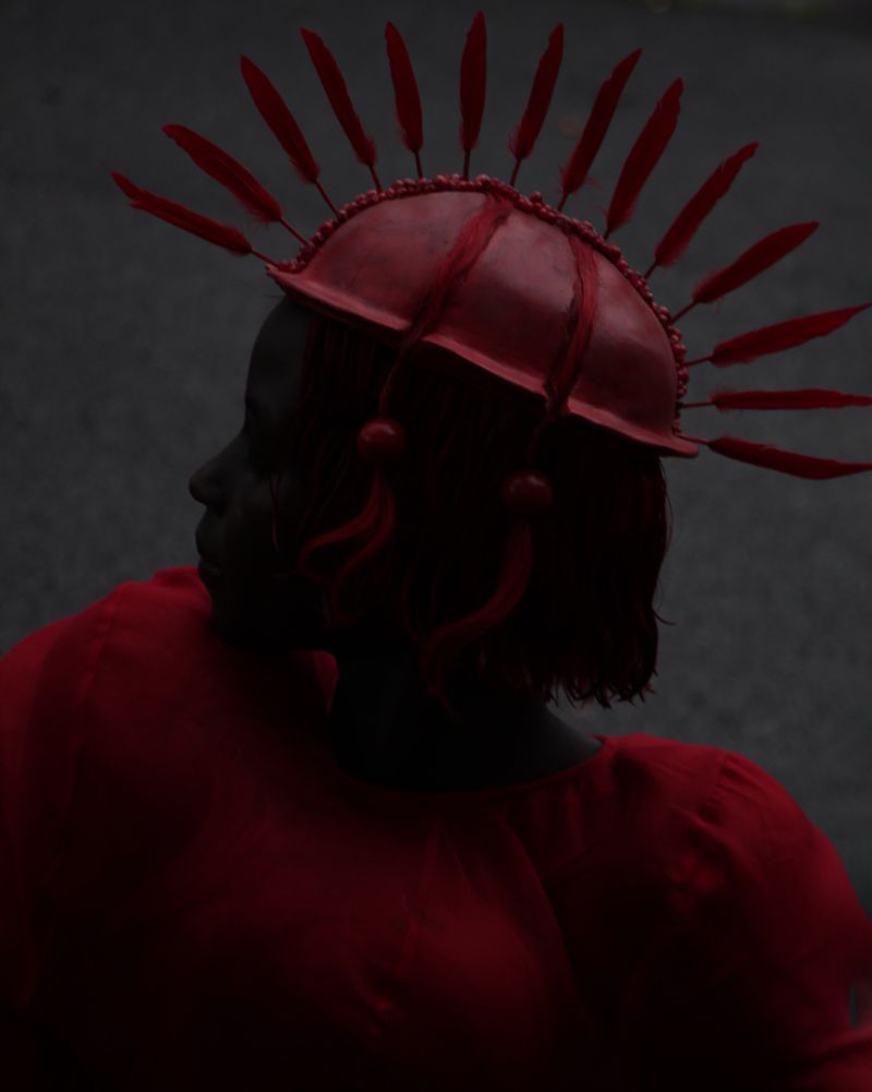 Woman in red with feather headdress in dark, moody setting. Shoulder-up studio portrait with dramatic lighting, side view.